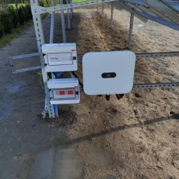 Instalacja fotowoltaiczna w trakcie montażu: widoczne panele słoneczne na metalowej konstrukcji, skrzynki elektryczne z otwartą obudową i inwerter na ziemi.