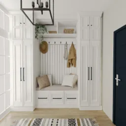 Biała zabudowa przedpokoju z siedziskiem, wieszakami i szafkami, uzupełniona o dekoracje jak kapelusz, roślina i dywanik. Ciemne drzwi po prawej.