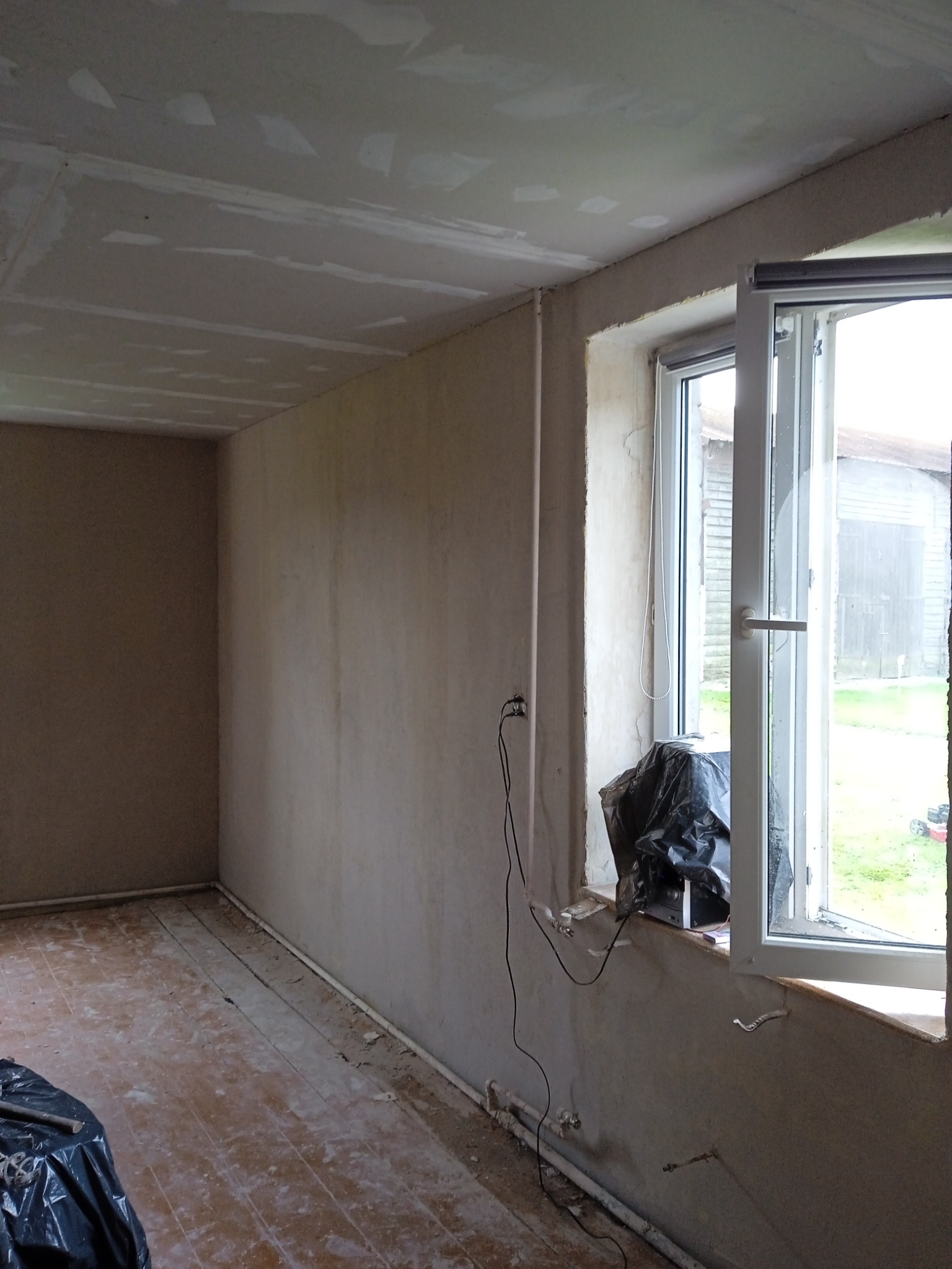 Pokój w trakcie prac wykończeniowych: ściany po szpachlowaniu, sufit z płyt karton-gips, okno otwarte, podłoga drewniana częściowo zakryta folią.