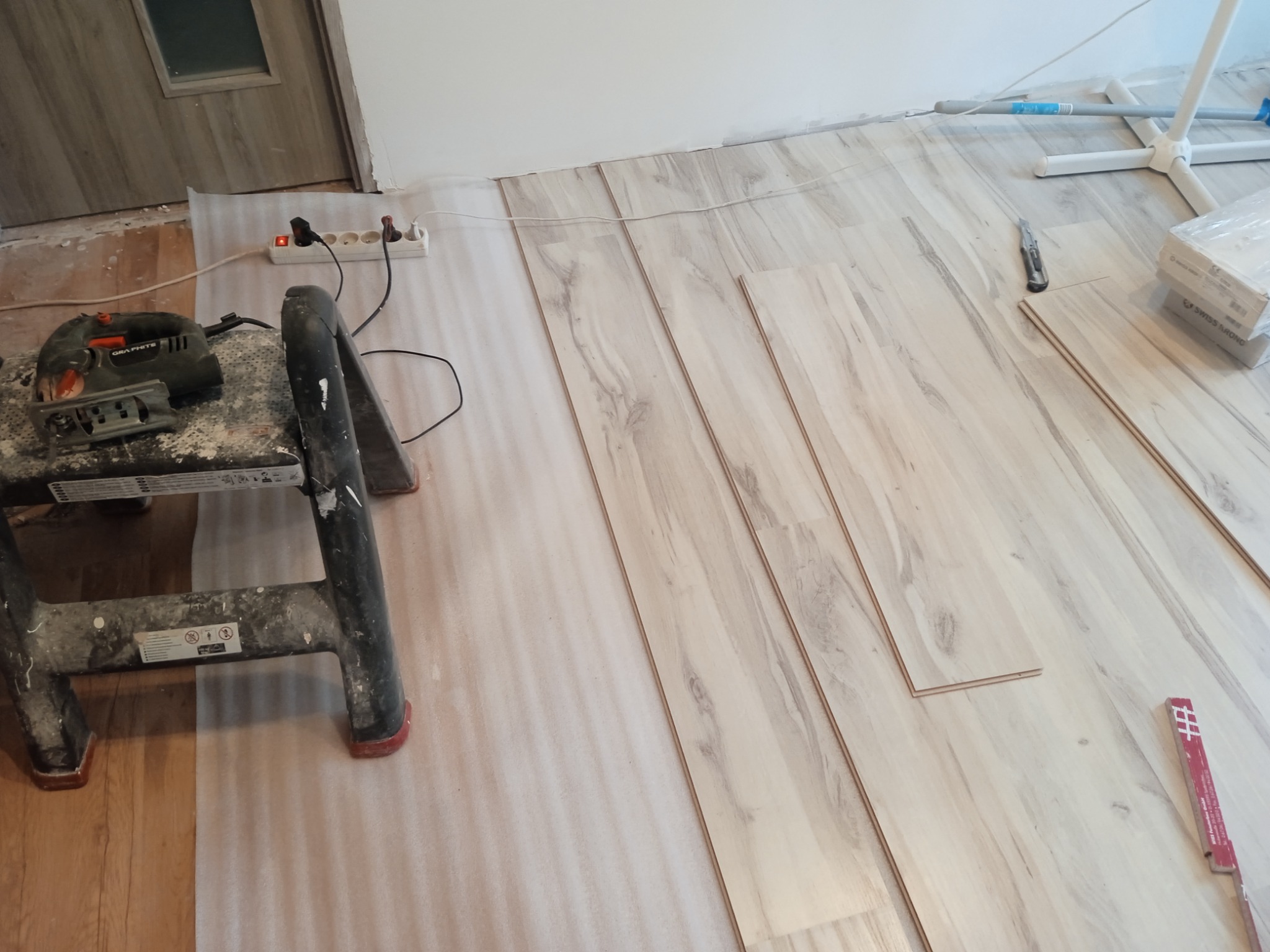 Fragment układania paneli podłogowych w jasnym odcieniu drewna, widoczna piła elektryczna na zabrudzonym stołku, nóż do cięcia paneli oraz rozłożona folia ochronna.