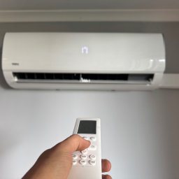 TECHCEL MARCEL ŻYDEK - Biały klimatyzator ścienny włączony pilotem. Na wyświetlaczu klimatyzatora widoczna temperatura 17 stopni. Pilot w dłoni, ustawienia temperatury.