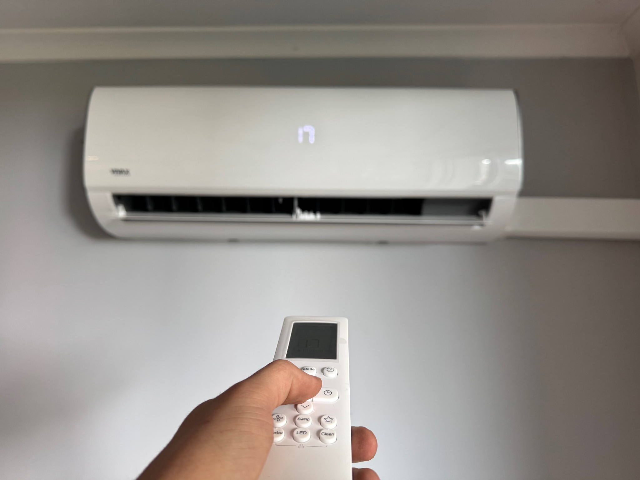 Biały klimatyzator ścienny włączony pilotem. Na wyświetlaczu klimatyzatora widoczna temperatura 17 stopni. Pilot w dłoni, ustawienia temperatury.