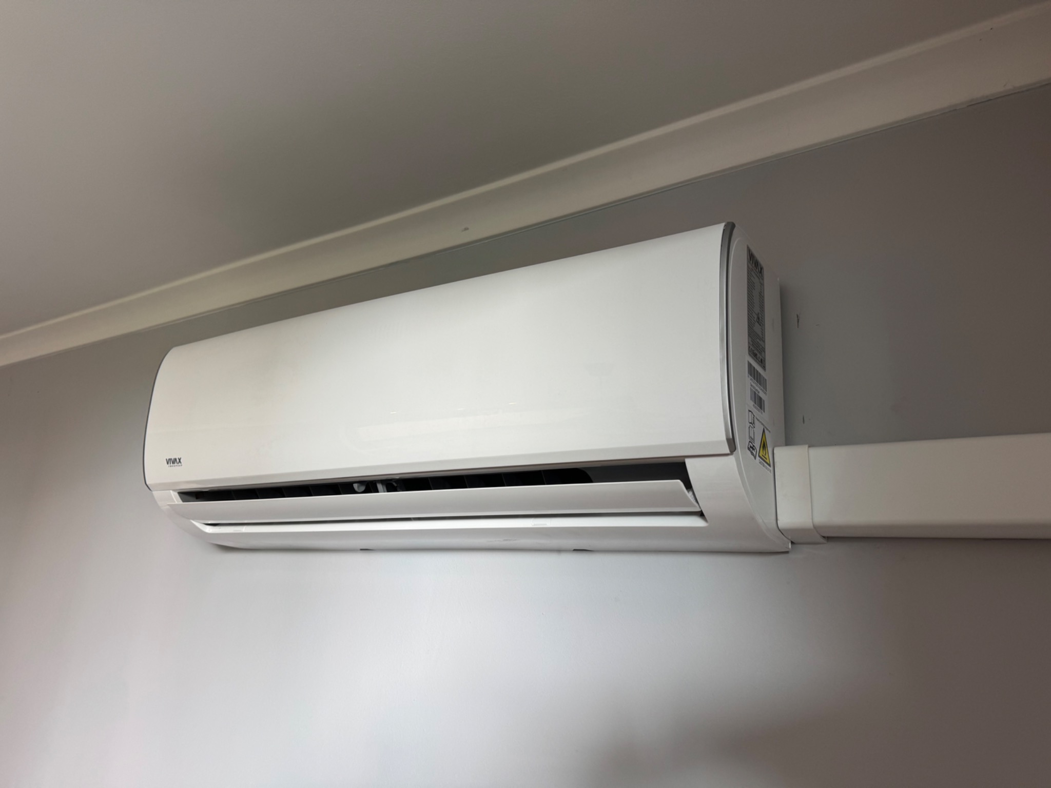 Biały klimatyzator VIVAX zamontowany na szarej ścianie, widoczne przewody w białej maskownicy. Minimalistyczny design, nowoczesne rozwiązanie dla domu lub biura.