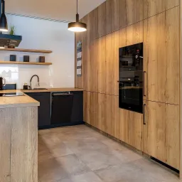 Nowoczesna kuchnia z drewnianymi frontami szafek, czarnymi akcentami i oświetleniem punktowym, widok na zabudowę piekarnika i mikrofalówki.