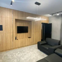 Salon z zabudową meblową z jasnego drewna, czarną sofą i oświetleniem LED, minimalistyczny design.