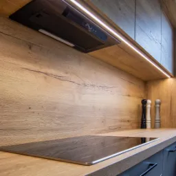 Fragment nowoczesnej kuchni z drewnianym blatem i panelem ściennym o wyraźnym usłojeniu, czarna płyta indukcyjna, okap podszafkowy z oświetleniem LED, młynki do przypraw i roślina w czerwonej...