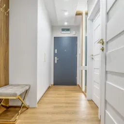 Nowoczesny przedpokój z drewnianą okładziną ścienną, złotymi haczykami i ławką z welurowym siedziskiem na złotym stelażu, szara ściana z szafą wnękową, drewniana podłoga, szare drzwi wejściowe...