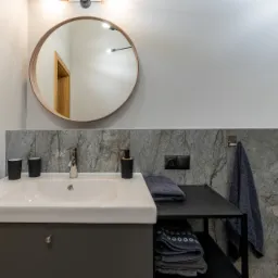 Nowoczesna łazienka z umywalką nablatową, okrągłym lustrem w drewnianej ramie, szarą armaturą i marmurowymi płytkami ściennymi.