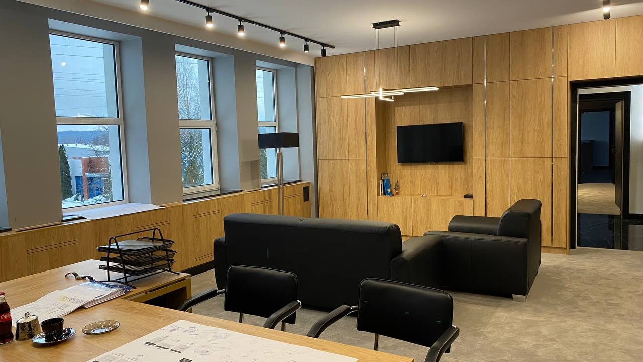 Nowoczesne biuro z drewnianą zabudową, czarnymi meblami wypoczynkowymi i dużymi oknami z widokiem na zimowy krajobraz, na biurku projekty i przybory.