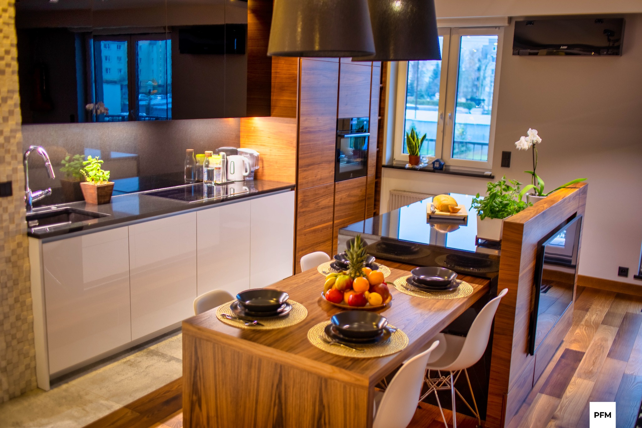 Nowoczesna kuchnia z drewnianymi i białymi szafkami, czarnym blatem i wyspą z drewnianym stołem oraz owocami w misie, widok na okno z roślinami.