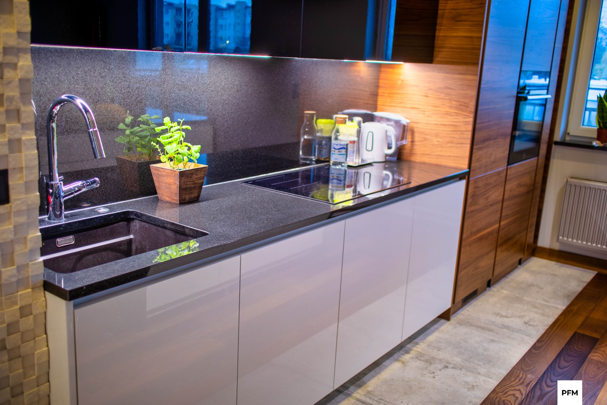 Nowoczesna kuchnia z ciemnym blatem granitowym, chromowaną baterią i drewnianymi akcentami, oświetlona listwą LED pod szafkami wiszącymi, z widocznym fragmentem podłogi z desek.