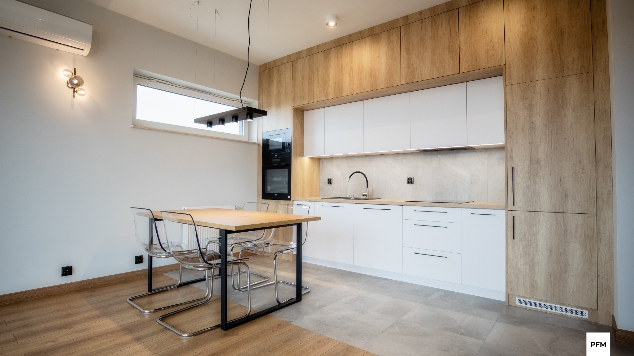 Nowoczesna kuchnia z białymi frontami szafek dolnych i górnymi szafkami w odcieniu jasnego drewna, czarny zlew i bateria, stół z drewnianym blatem i przezroczystymi krzesłami.