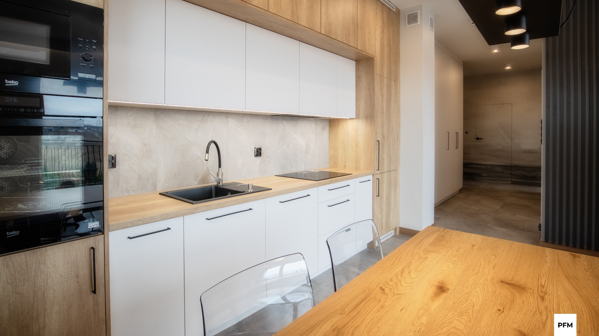 Nowoczesna kuchnia z białymi szafkami, drewnianym blatem i czarnym zlewozmywakiem, widok z perspektywy stołu z przezroczystymi krzesłami, w tle widoczny korytarz z szafą w zabudowie.