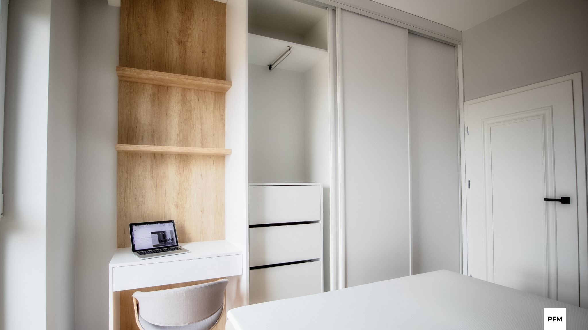 Nowoczesny pokój z białą szafą przesuwną, biurkiem z drewnianymi półkami i laptopem, oraz białymi drzwiami z czarną klamką.