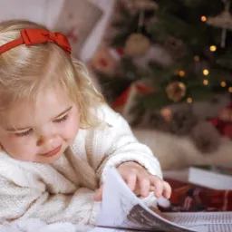 Mała dziewczynka z blond włosami i czerwoną kokardą na głowie, ubrana w biały sweter, leży na podłodze i ogląda książkę na tle choinki z delikatnymi światełkami.