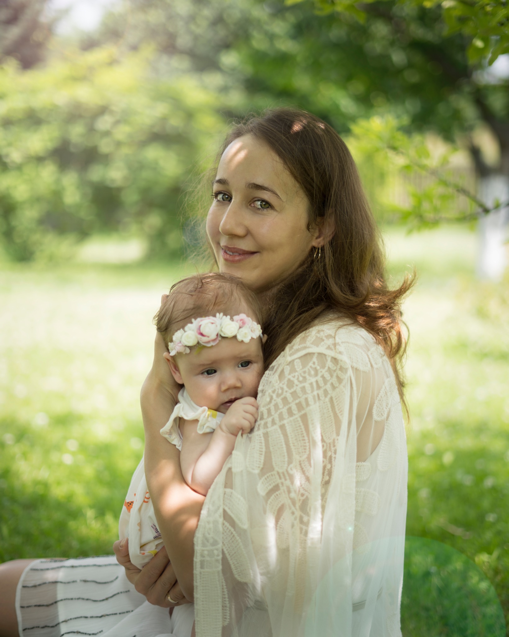 Portret matki trzymającej na rękach noworodka z kwiatowym wiankiem na głowie, na tle zielonego ogrodu w słoneczny dzień.