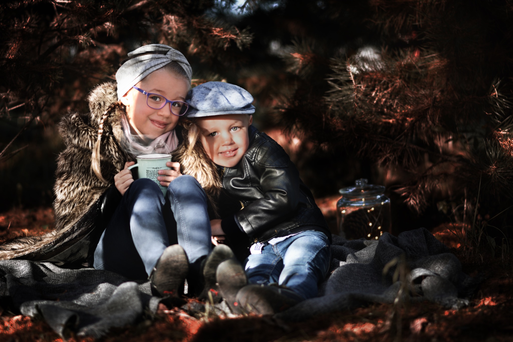 Dwoje dzieci, dziewczynka w okularach trzymająca kubek i chłopiec w czapce i kurtce, siedzą na kocu w jesiennej scenerii z jarzącym się słoikiem w tle.