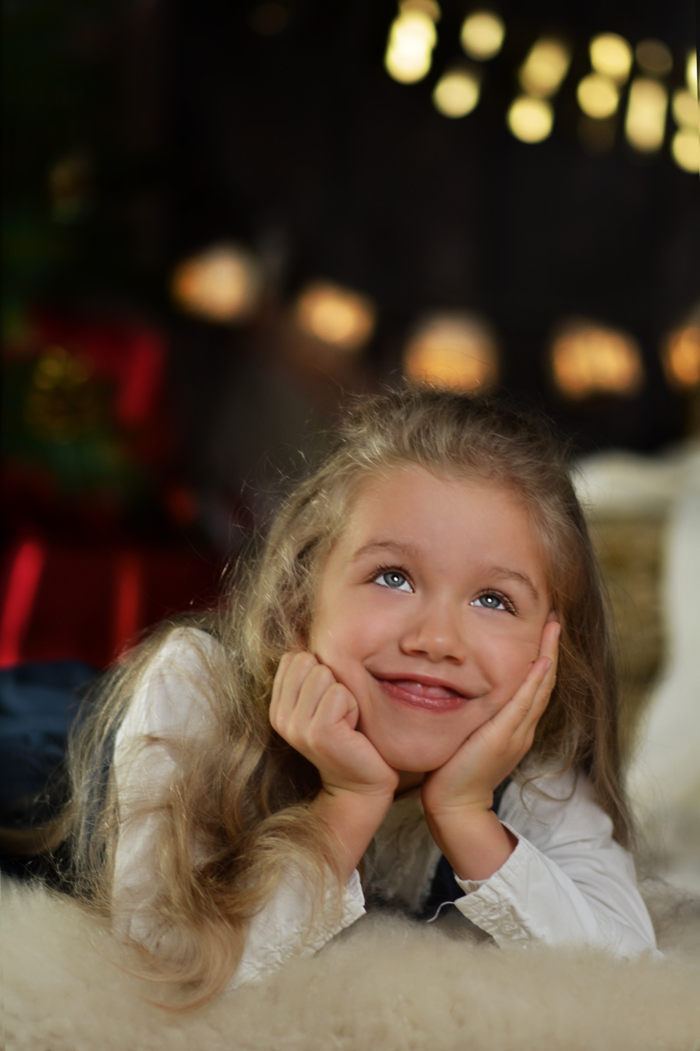 Uśmiechnięta dziewczynka z blond włosami leży na puszystym dywanie, podpierając brodę dłońmi i spoglądając w górę, w tle rozmyte światełka.