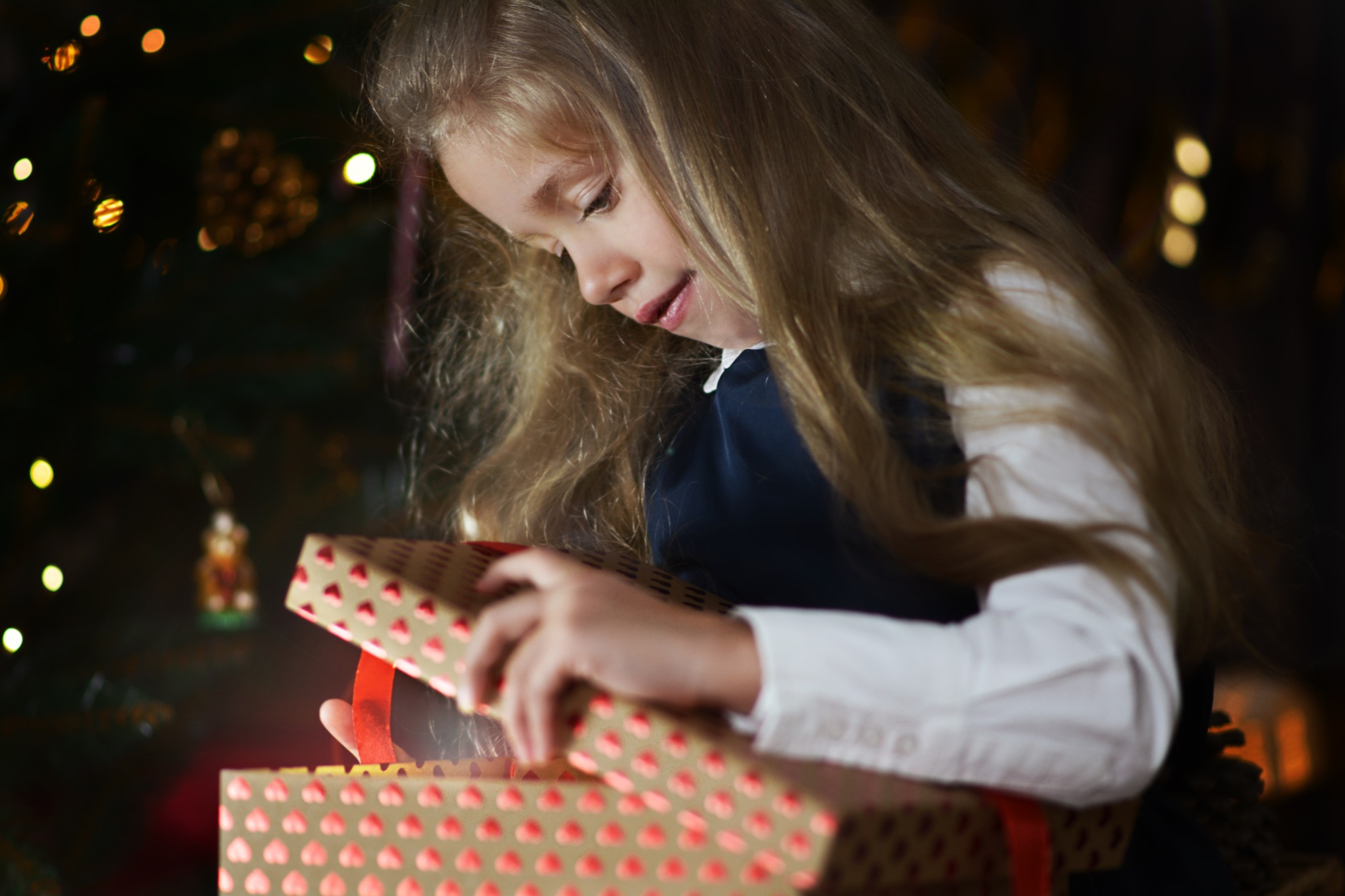 Dziewczynka z długimi blond włosami otwiera pudełko prezentowe z czerwonymi serduszkami na tle choinki z delikatnym oświetleniem, emanującym ciepłym blaskiem.