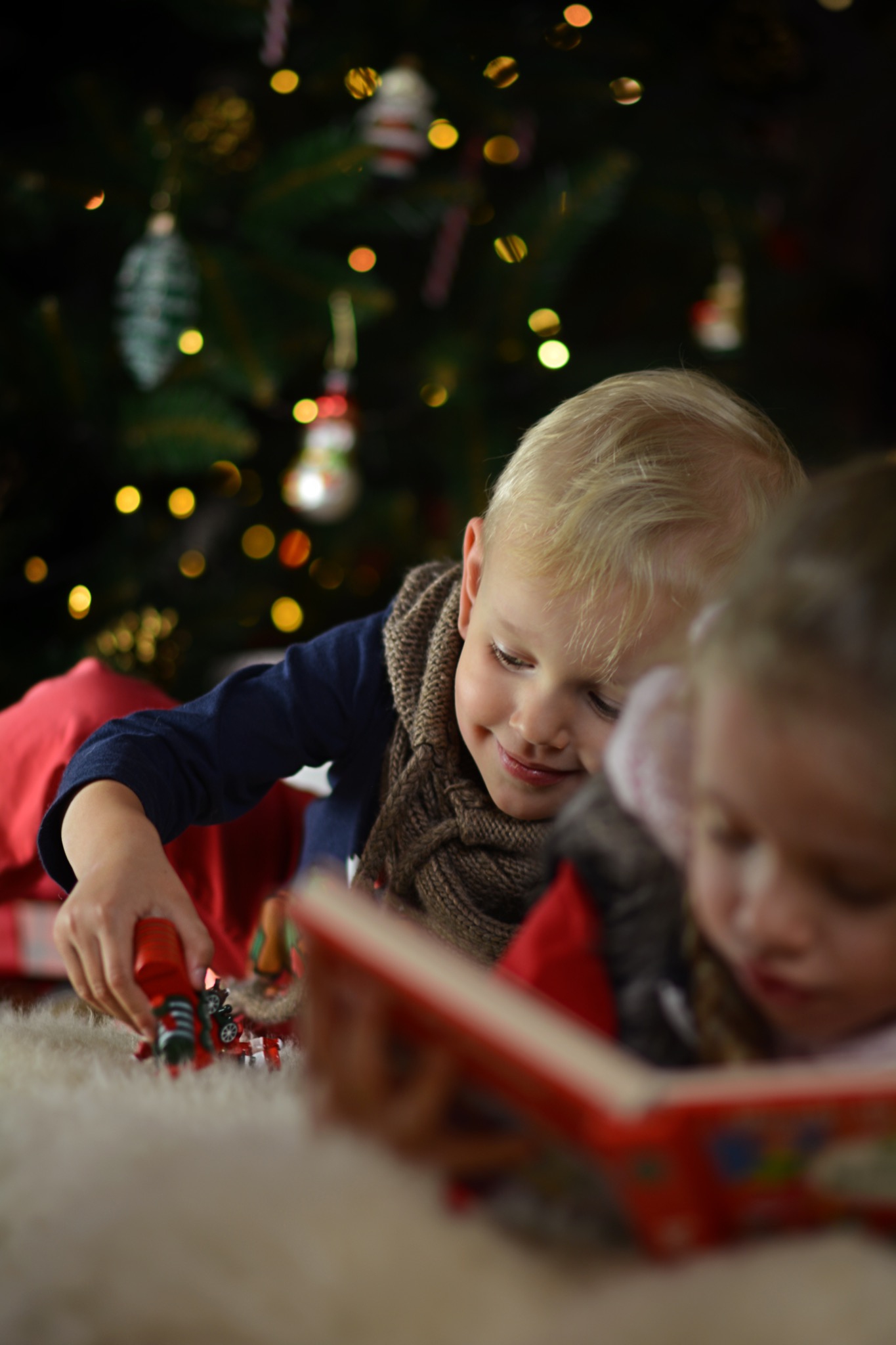 Dwoje dzieci leżących na dywanie z choinką w tle: chłopiec bawi się czerwoną zabawką, a dziewczynka czyta książkę.