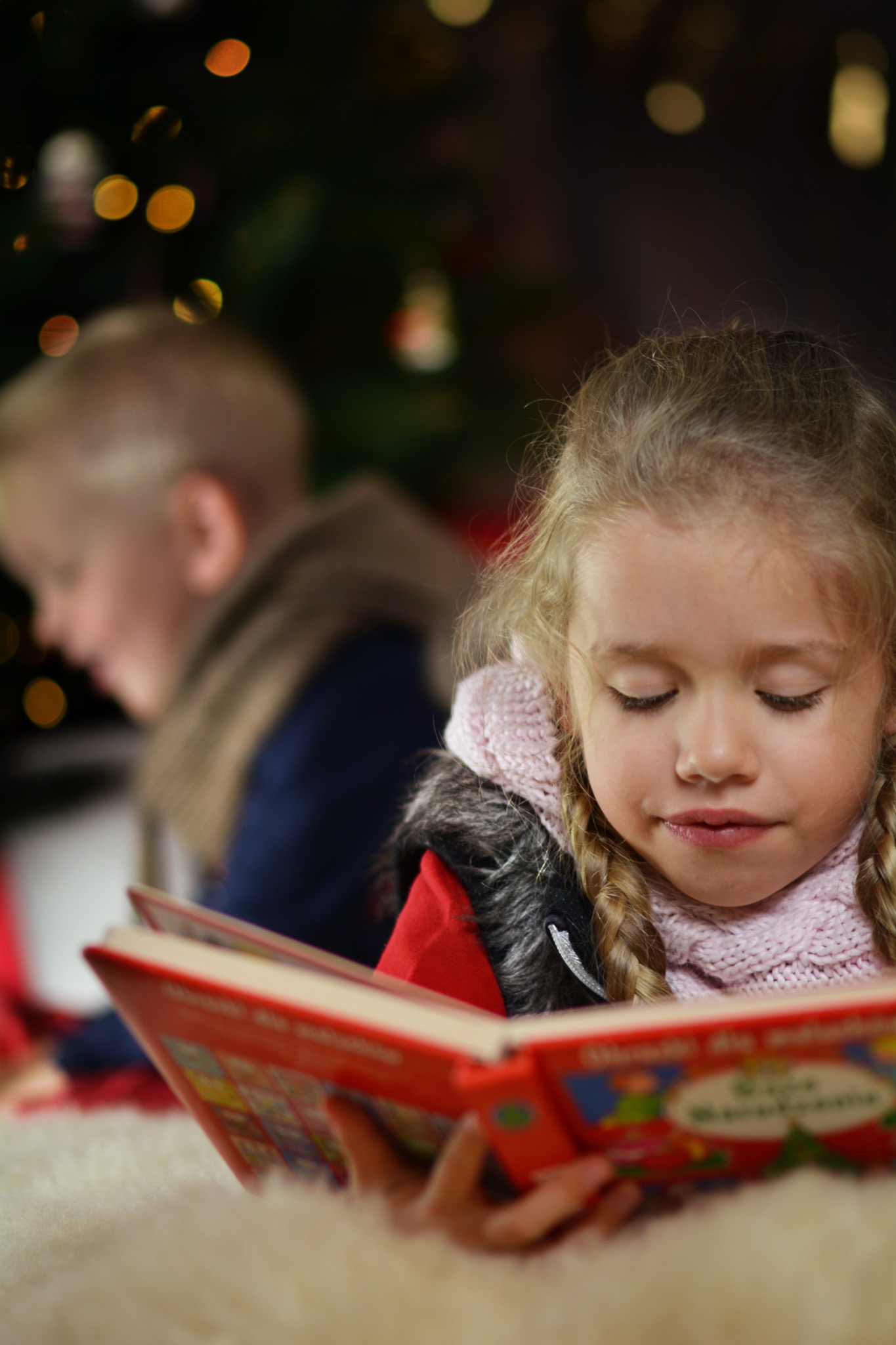 Dziewczynka z warkoczami czyta książkę z kolorowymi ilustracjami na tle rozświetlonej choinki, w tle widoczny chłopiec.