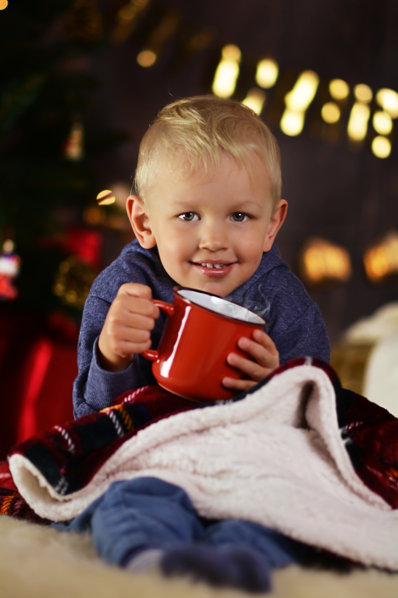 Uśmiechnięty blond chłopiec trzymający czerwony kubek z parującym napojem, otulony w koc w kratę na tle rozmytych światełek.