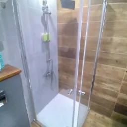 Kabina prysznicowa z deszczownicą i szarą ścianą, wykończona drewnopodobnymi płytkami, z zieloną gąbką na słuchawce prysznicowej.