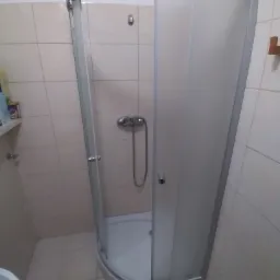 Kabina prysznicowa narożna z matową szybą, zamontowana w łazience wyłożonej jasnymi, kwadratowymi płytkami. Widoczny srebrny panel prysznicowy z wężem i słuchawką.