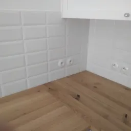 Białe płytki typu metro z widocznymi gniazdkami elektrycznymi nad drewnianym blatem kuchennym i fragmentem białej szafki wiszącej.