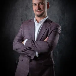 Portret mężczyzny w bordowym garniturze w kratę, z rękami skrzyżowanymi na piersiach, na ciemnym, teksturowanym tle.