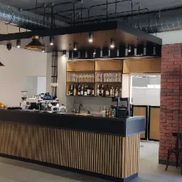 Wnętrze lokalu gastronomicznego z piecem do pizzy, barem z ekspresami do kawy i regałami z napojami, oświetlone wiszącymi lampami w stylu industrialnym.