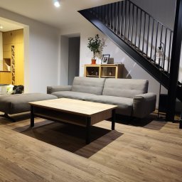 HIMEBLE SPÓŁKA Z OGRANICZONĄ ODPOWIEDZIALNOŚCIĄ - Nowoczesny salon z szarą sofą, drewnianym stolikiem kawowym i czarnymi schodami z metalową balustradą, uzupełniony drewnianą podłogą i widoczną kuchnią w tle.