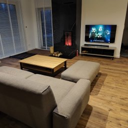 HIMEBLE SPÓŁKA Z OGRANICZONĄ ODPOWIEDZIALNOŚCIĄ - Nowoczesny salon z drewnianą podłogą, szarą sofą, stolikiem kawowym, czarnym kominkiem z marmurowym wykończeniem i telewizorem wyświetlającym zespół Queen.