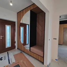 Wnęka z siedziskiem obita tkaniną w geometryczny wzór, obudowana lamelami i lustrem w kształcie koła, widok na drzwi wejściowe i wnętrze domu, podłoga częściowo zakryta kartonami.