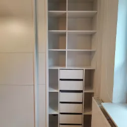 Biała szafa wnękowa z przesuwnymi drzwiami i otwartymi półkami z szufladami, widok na wnętrze szafy, podłoga w brązowo-białą szachownicę.