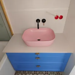 Łazienka z różową umywalką nablatową, czarną armaturą, niebieską szafką z ozdobnymi uchwytami i wzorzystą podłogą, widoczne niedokończone podłączenie elektryczne.