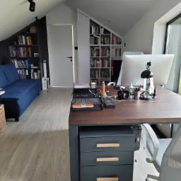 Biuro w domu na poddaszu ze skośnymi ścianami, z biurkiem z szufladami, komputerem iMac, lampką, sofą i regałami na książki.