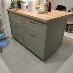 HIMEBLE SPÓŁKA Z OGRANICZONĄ ODPOWIEDZIALNOŚCIĄ - Wyspa kuchenna z drewnianym blatem i oliwkowymi szafkami, złote uchwyty, na blacie akcesoria kuchenne, widok z boku, szare płytki na podłodze.