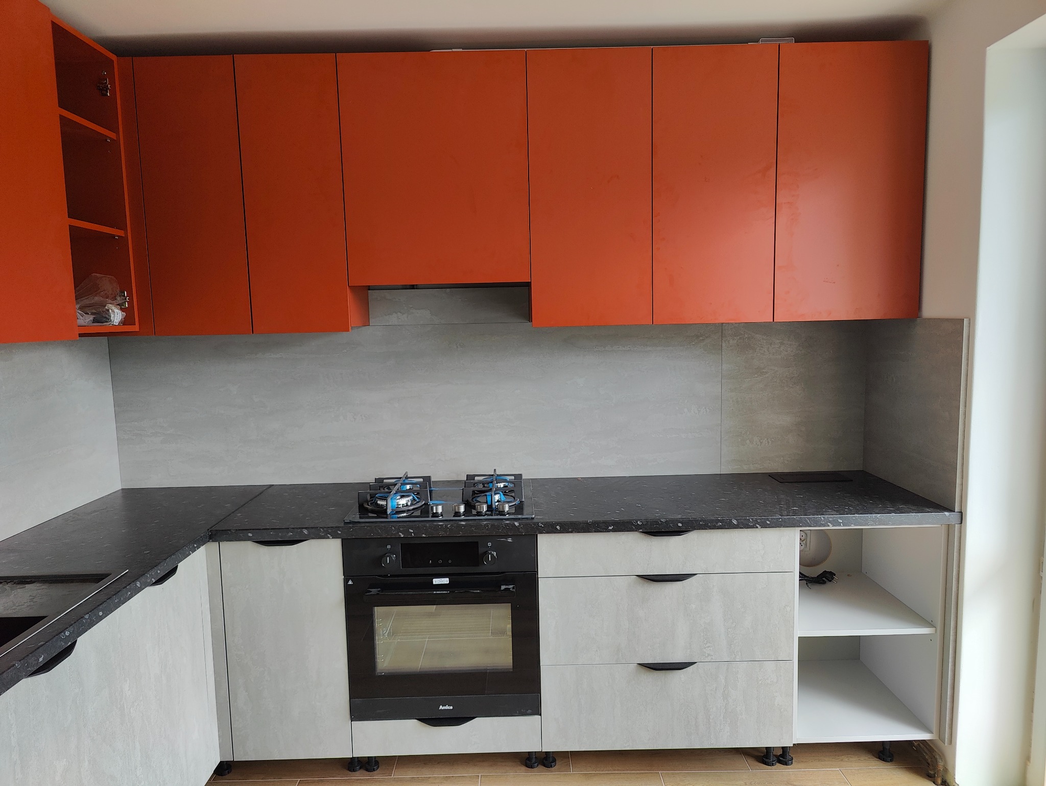 Nowoczesna kuchnia z pomarańczowymi szafkami wiszącymi i szarymi szafkami dolnymi z czarnym blatem, płyta gazowa z niebieskim płomieniem, piekarnik Amica.