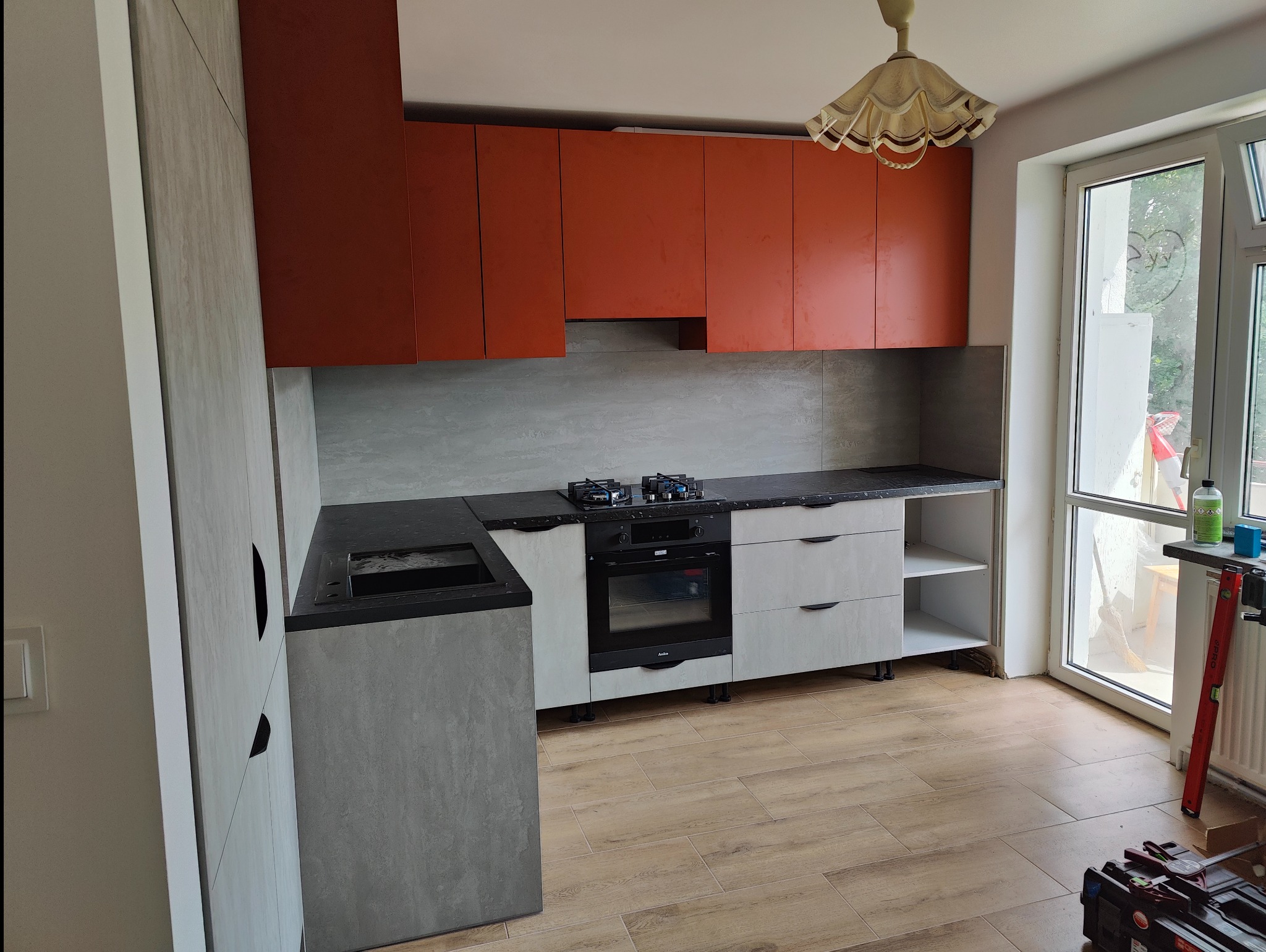 Nowoczesna kuchnia z pomarańczowymi szafkami górnymi i szarymi dolnymi, czarny blat, płyta gazowa i piekarnik, drewnopodobne płytki na podłodze, widoczne narzędzia i chemia gospodarcza sugerują...