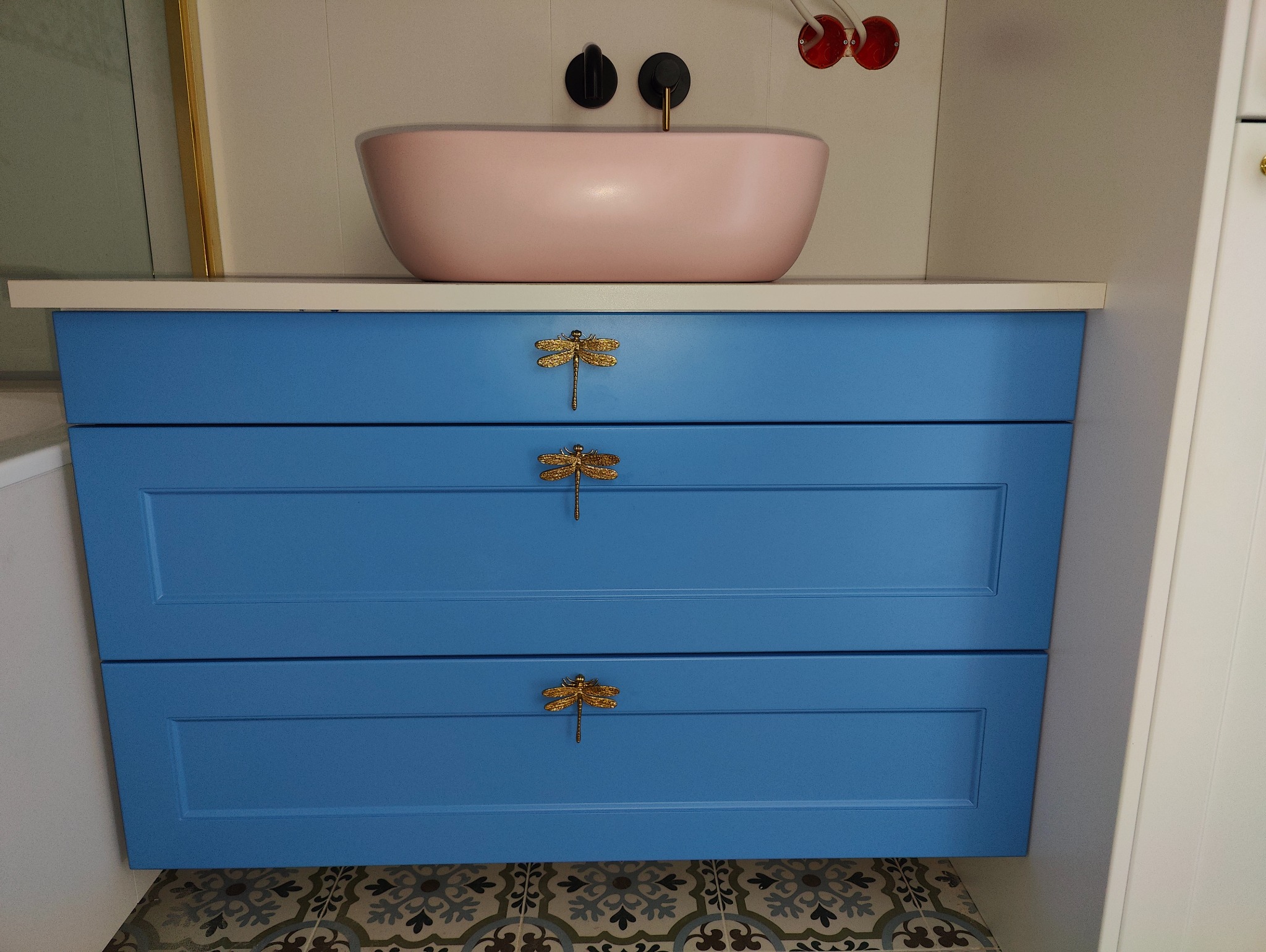 Niebieska szafka łazienkowa z trzema szufladami i złotymi uchwytami w kształcie ważek, nad nią różowa umywalka nablatowa, widoczne fragmenty instalacji wodnej i dekoracyjna podłoga.
