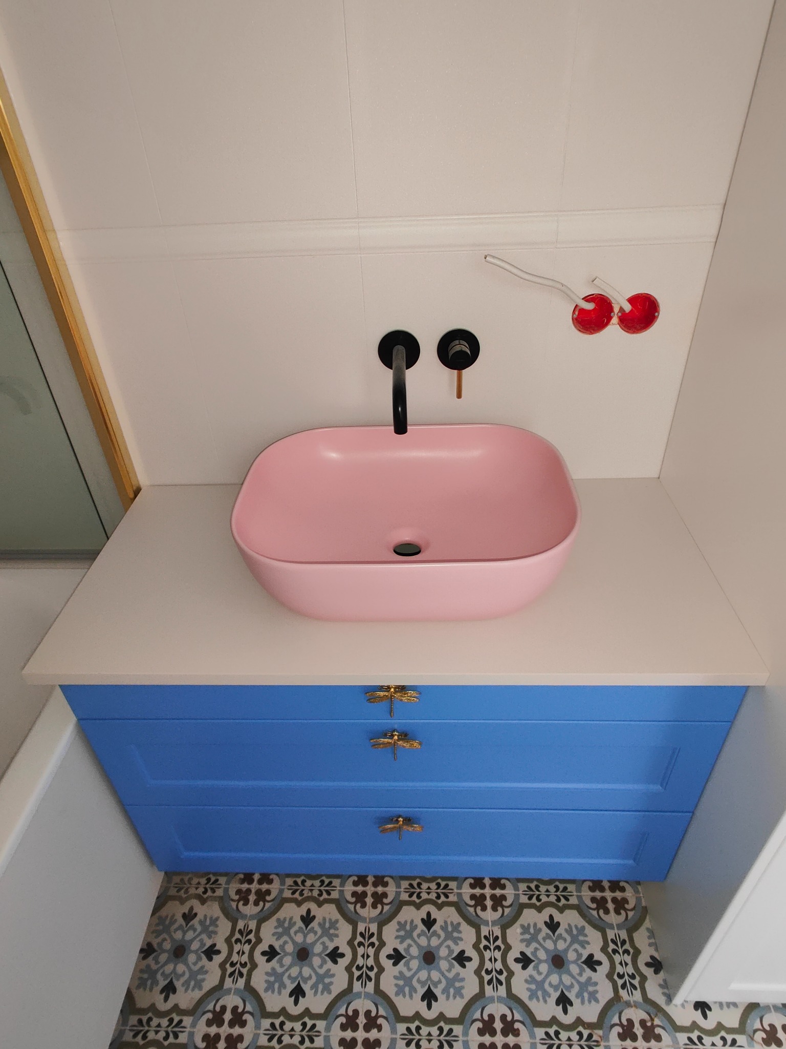 Łazienka z różową umywalką nablatową, czarną armaturą, niebieską szafką z ozdobnymi uchwytami i wzorzystą podłogą, widoczne niedokończone podłączenie elektryczne.
