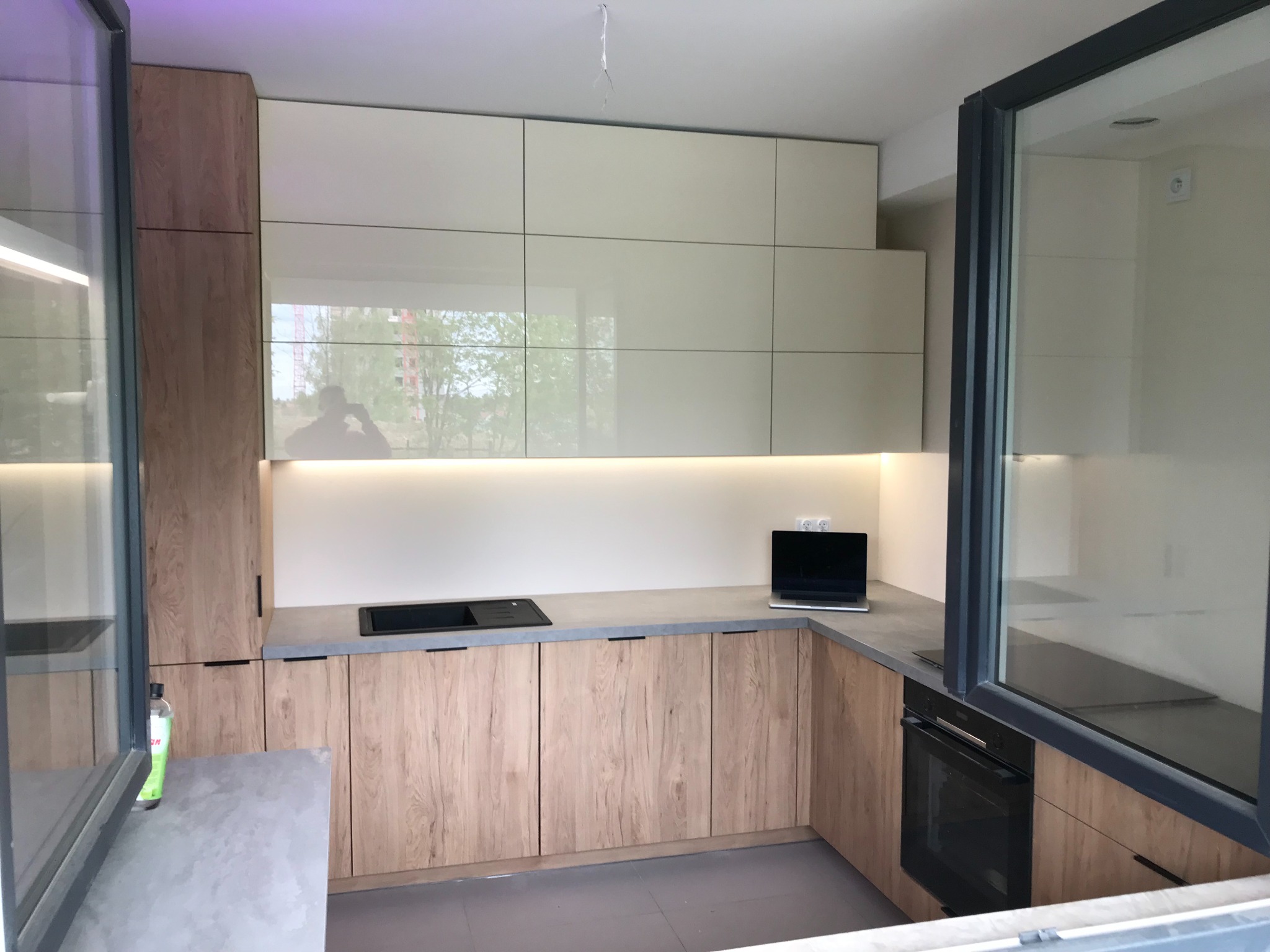 Nowoczesna kuchnia z drewnianymi frontami szafek dolnych i białymi, błyszczącymi szafkami górnymi, szary blat, czarny zlew i piekarnik, laptop na blacie, duże okno z czarną ramą, widoczny fragment...