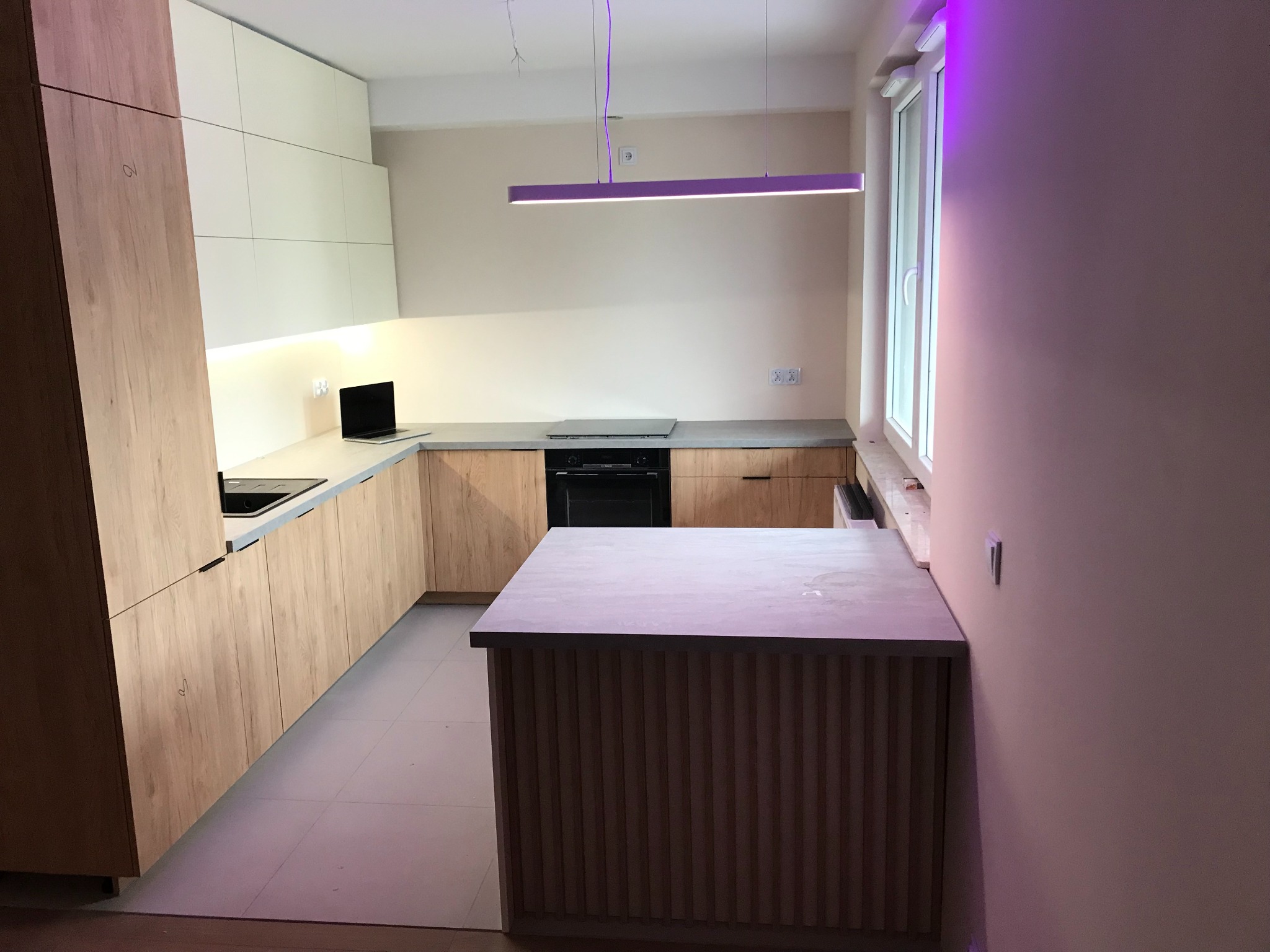 Nowoczesna kuchnia z drewnianymi frontami, szarym blatem i wyspą z ryflowanym wykończeniem, oświetlona podłużną lampą LED w kolorze fioletowym.