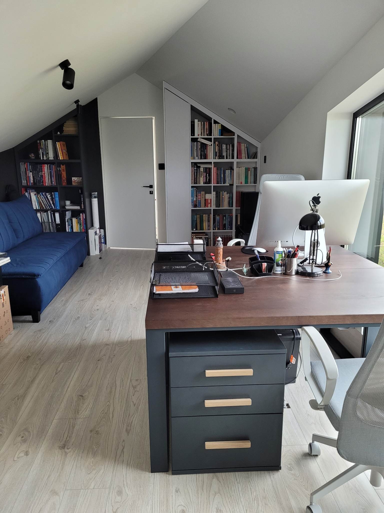 Biuro w domu na poddaszu ze skośnymi ścianami, z biurkiem z szufladami, komputerem iMac, lampką, sofą i regałami na książki.