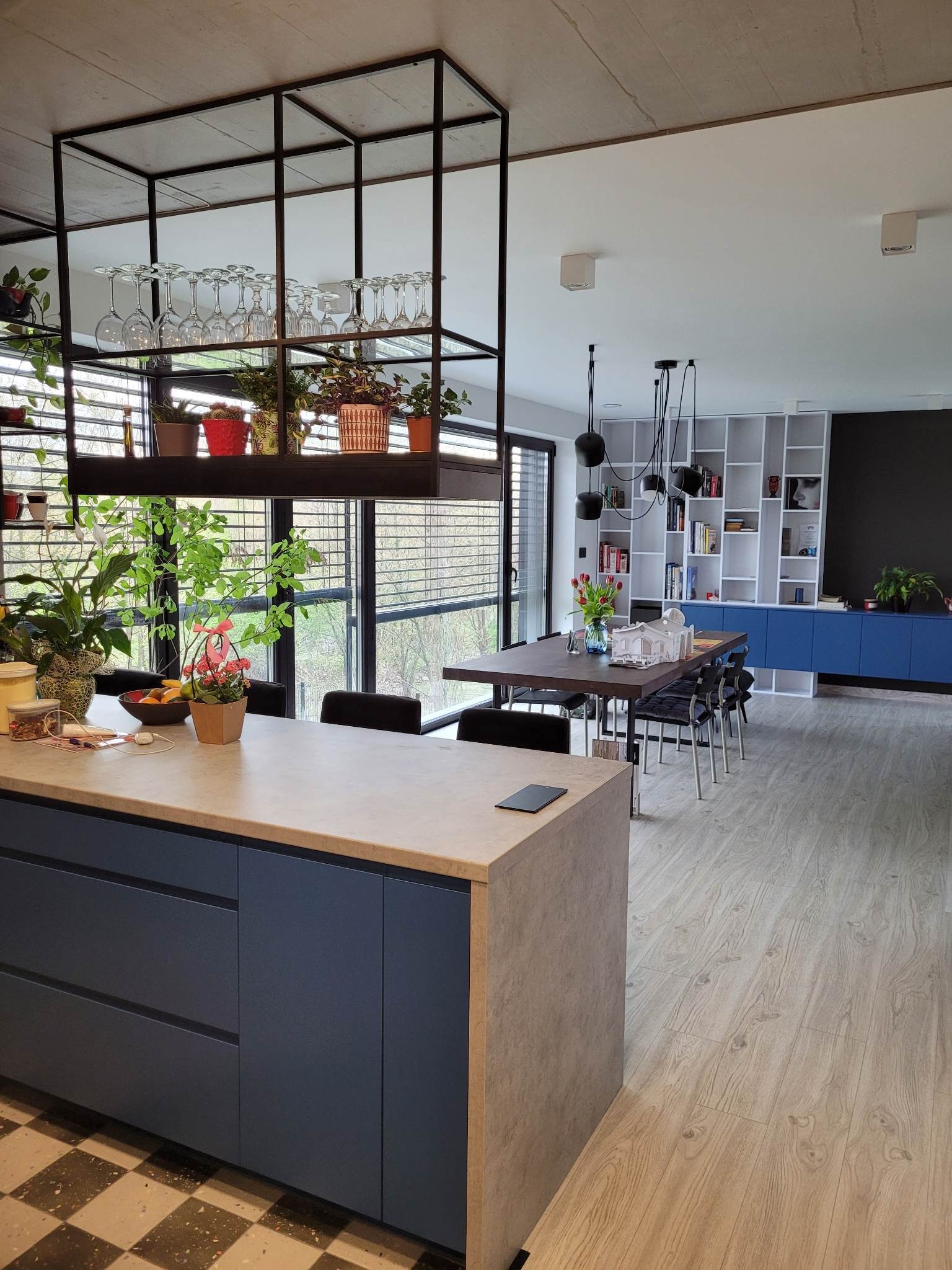 Nowoczesna kuchnia z wyspą i metalową konstrukcją nad nią, na której znajdują się kieliszki i rośliny doniczkowe, widok na jadalnię z niebieską zabudową i czarną ścianą.