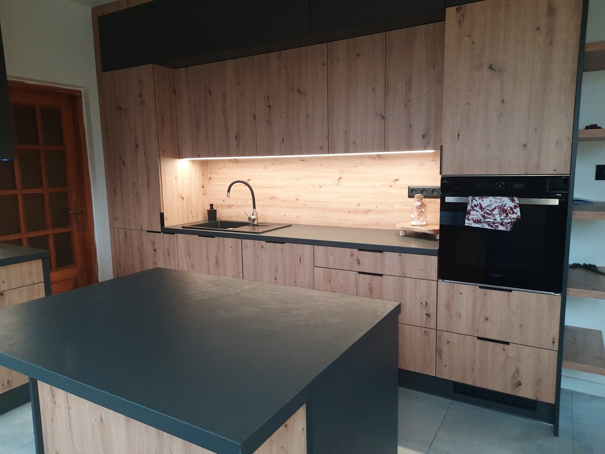 Nowoczesna kuchnia z drewnopodobnymi frontami i czarnymi blatami, oświetlenie LED pod szafkami wiszącymi, wyspa kuchenna z czarnym blatem, piekarnik w zabudowie.