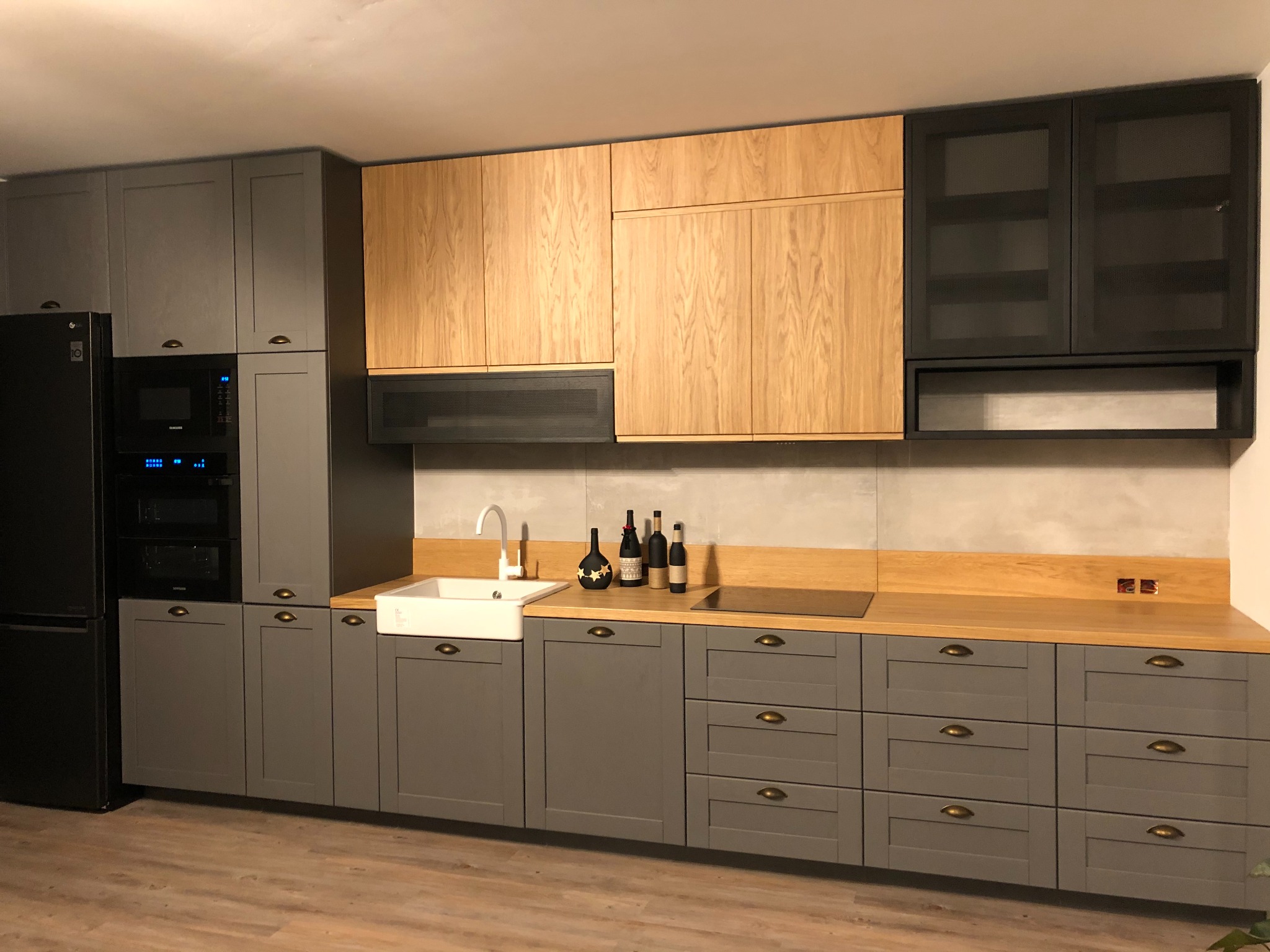 Nowoczesna kuchnia z szarymi szafkami dolnymi i drewnianymi górnymi, z ceramicznym zlewem farmerskim i czarną lodówką.