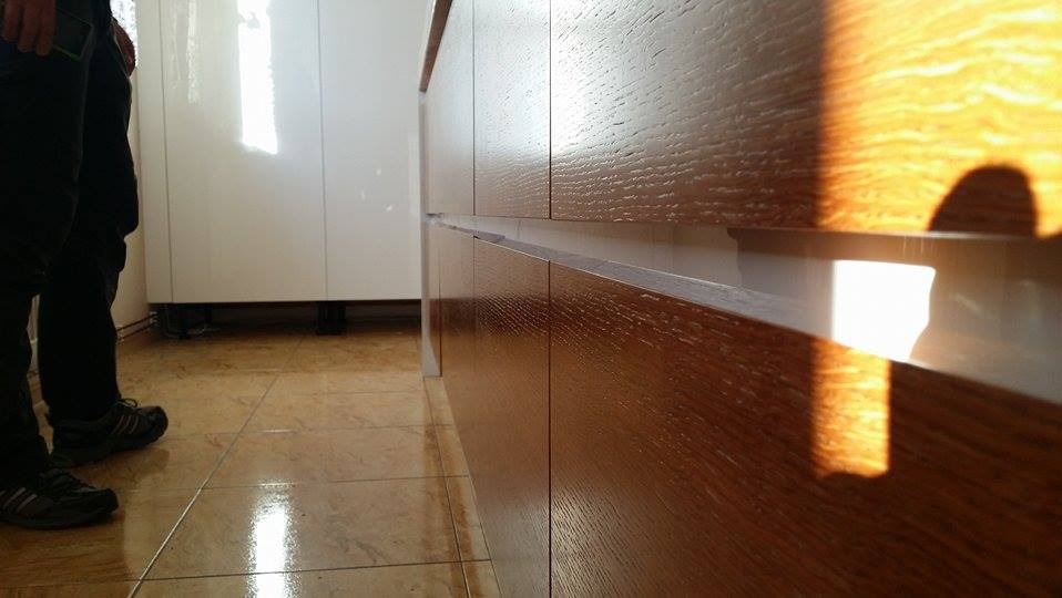 Fragment zabudowy kuchennej z drewnianymi frontami, widoczny fragment podłogi z płytek i noga osoby stojącej obok.