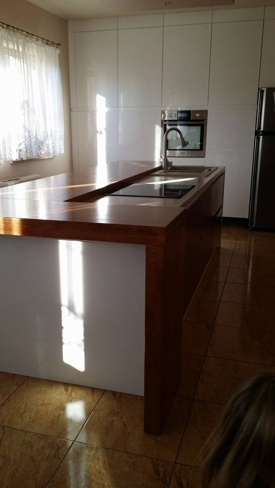 Nowoczesna kuchnia z białą zabudową i drewnianym blatem wyspowym z wbudowaną płytą grzewczą i zlewem, oświetlona naturalnym światłem wpadającym przez okno.